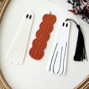 May include: Three bookmarks arranged vertically. One white ghost bookmark with a subtle floral pattern, one stack of five orange pumpkins with a gold stem, and one white ghost bookmark with black line details and a black tassel attached at the top.