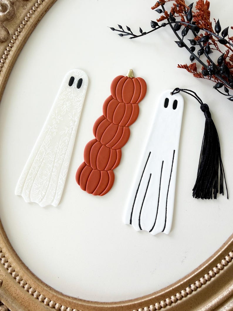 May include: Three bookmarks arranged vertically. One white ghost bookmark with a subtle floral pattern, one stack of five orange pumpkins with a gold stem, and one white ghost bookmark with black line details and a black tassel attached at the top.
