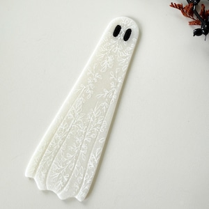 May include: A flat, ghost-shaped bookmark made of translucent white material. The surface features a delicate, white line-art pattern of various leaves and floral stems. Two small, solid black oval shapes serve as eyes near the rounded top.