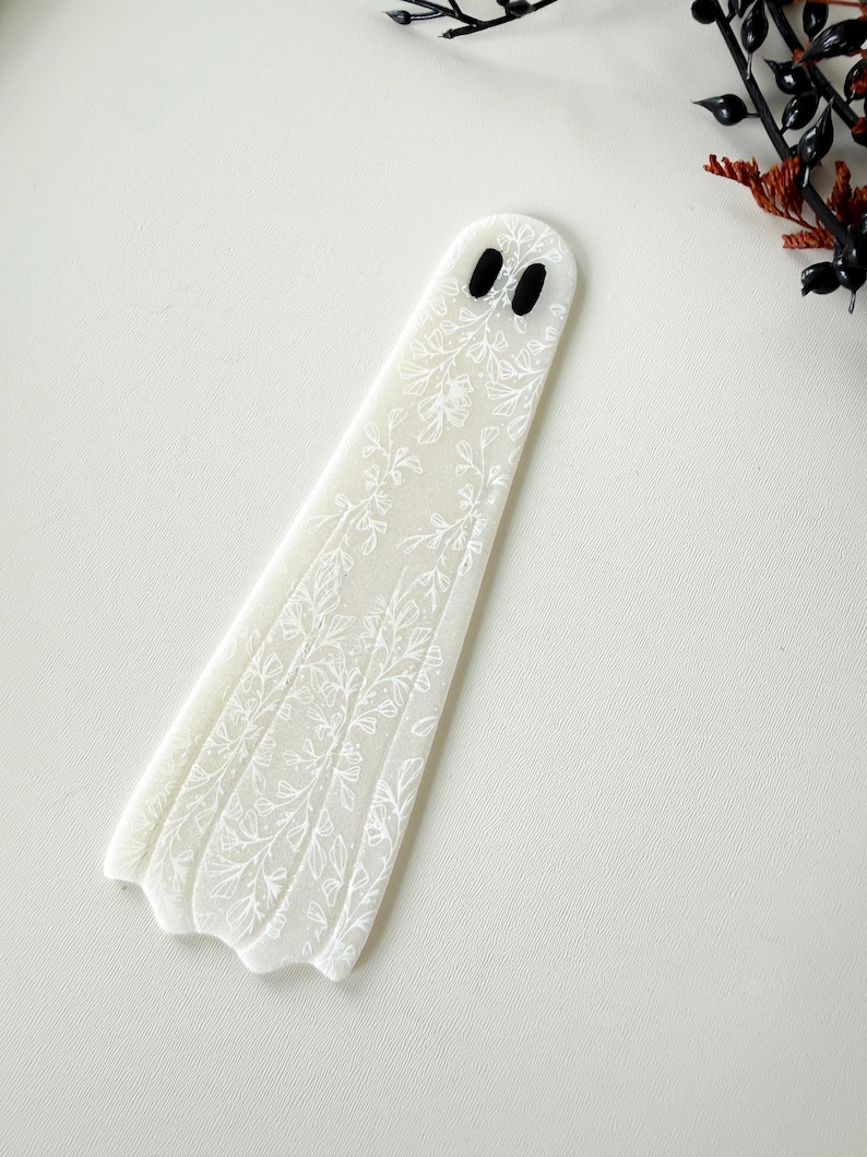 May include: A flat, ghost-shaped bookmark made of translucent white material. The surface features a delicate, white line-art pattern of various leaves and floral stems. Two small, solid black oval shapes serve as eyes near the rounded top.