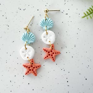 May include: Dangle earrings featuring a beach theme. Each earring has a gold-colored post and is composed of a light blue seashell, a white sand dollar, and a coral-colored starfish. The starfish has small, dark red dots.
