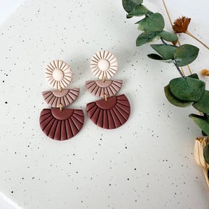 May include: A pair of statement earrings with three tiers. The top tier is a white circle with a textured design. The middle tier is a light pink half-circle with a textured design. The bottom tier is a dark red half-circle with a textured design.