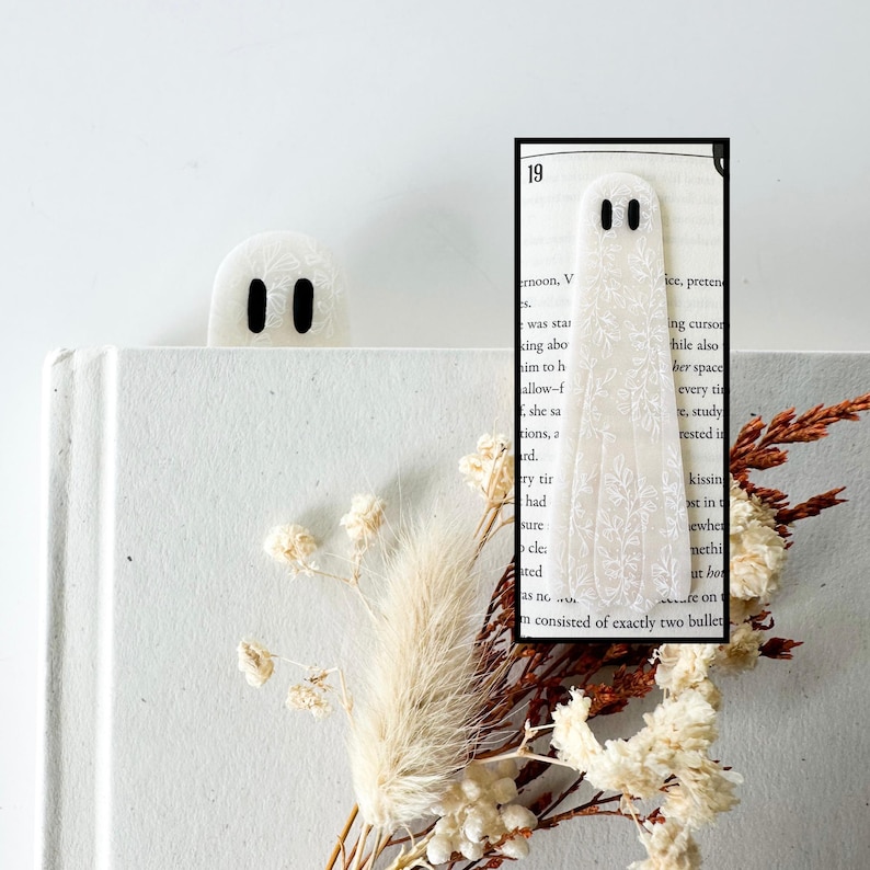 May include: A white ghost-shaped bookmark featuring a delicate white floral line art pattern and two black oval eyes. The bookmark is shown partially tucked into a book page, with a second ghost bookmark visible in the background.
