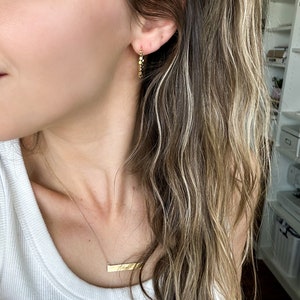 May include: A woman wearing a white vest top with a gold necklace featuring a horizontal bar pendant engraved with the word "fine". She is also wearing gold hoop earrings.