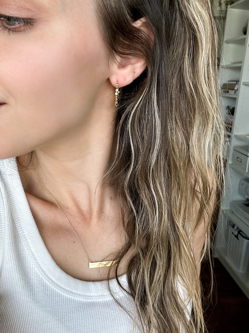 May include: A woman wearing a white vest top with a gold necklace featuring a horizontal bar pendant engraved with the word "fine". She is also wearing gold hoop earrings.