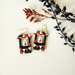 May include: A pair of dangle earrings featuring a miniature market stall design. Each stall is black with a red and white striped awning, a white ghost figure holding a small orange pumpkin, and orange triangular bunting along the base.