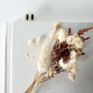 May include: A white ghost-shaped bookmark with two black oval eyes peeking out from the top of a light gray book. A small bouquet of dried cream-colored flowers and brown foliage is arranged on the front cover of the book.