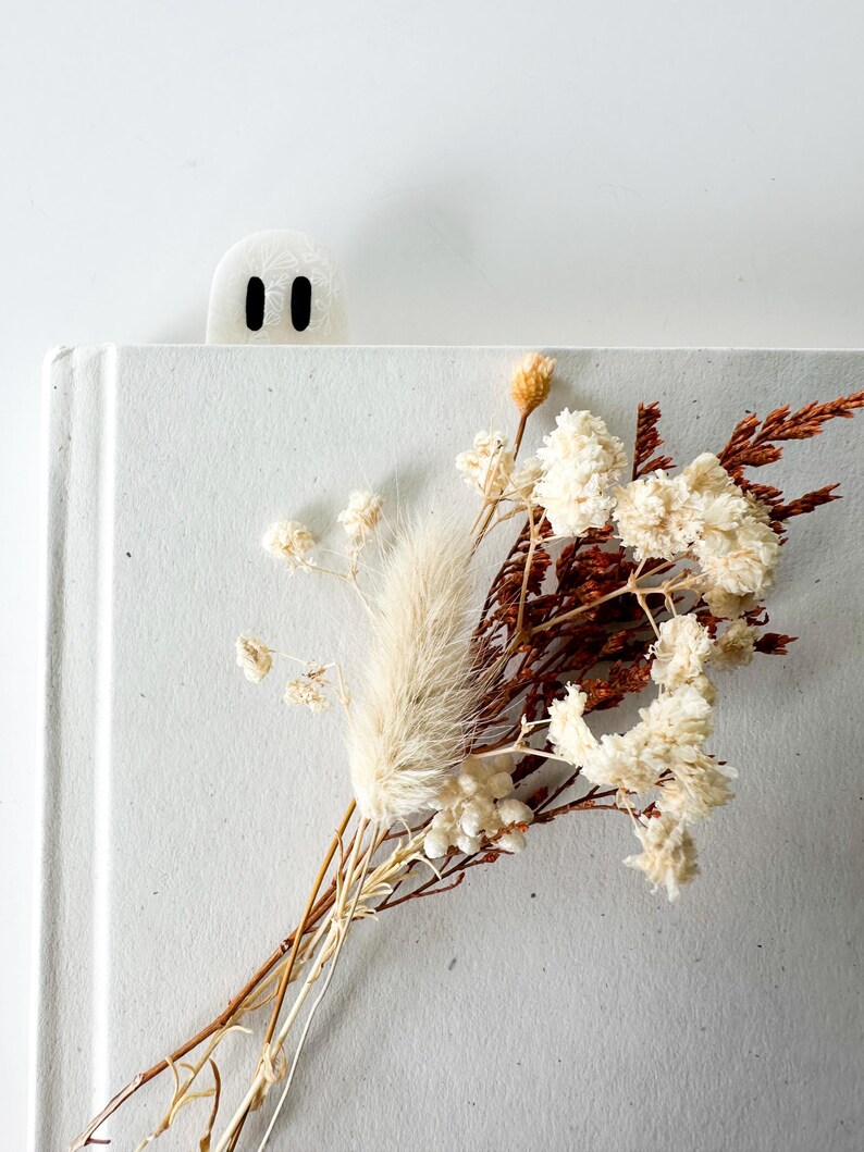 May include: A white ghost-shaped bookmark with two black oval eyes peeking out from the top of a light gray book. A small bouquet of dried cream-colored flowers and brown foliage is arranged on the front cover of the book.