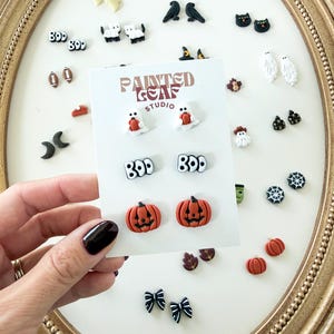 May include: A collection of small Halloween-themed stud earrings displayed on a white card labeled Painted Leaf Studio. The card features three pairs of earrings: white ghosts holding orange pumpkins, black and white text reading BOO, and orange jack-o'-lanterns.