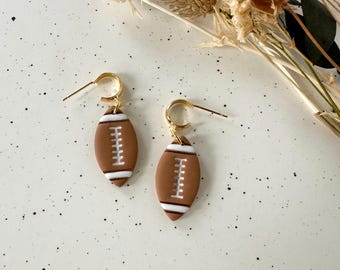 May include: A pair of earrings featuring a football design. The earrings are brown with white stitching and accents, and are suspended from gold-colored hoops. The earrings are displayed on a speckled white surface.