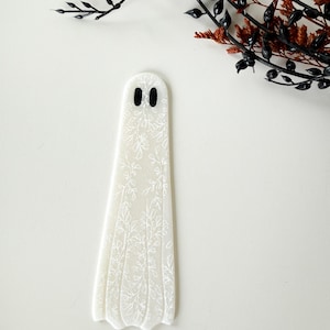 May include: A flat, ghost-shaped bookmark made of translucent white material. The surface features a delicate, white, etched floral pattern covering the entire body. Two small, solid black oval shapes serve as eyes near the top of the ghost figure.