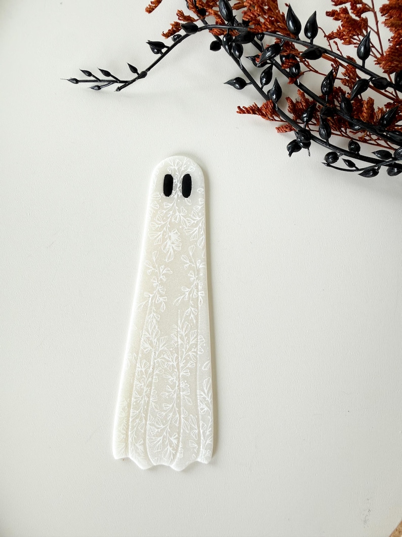 May include: A flat, ghost-shaped bookmark made of translucent white material. The surface features a delicate, white, etched floral pattern covering the entire body. Two small, solid black oval shapes serve as eyes near the top of the ghost figure.
