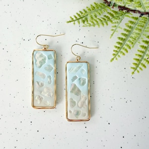 May include: A pair of rectangular earrings with a gold-colored frame. The earrings feature a light blue background with embedded white and light green stone-like shapes. The earrings are hanging from gold-colored ear wires.