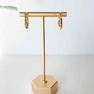 May include: Gold hoop earrings with a textured, beaded design. The earrings are displayed on a gold stand.