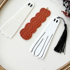 May include: Three bookmarks including a white ghost with a floral pattern, a stack of orange pumpkins, and a white ghost with black line details and a black tassel. Each bookmark features two black oval eyes at the top for a ghost-like appearance.