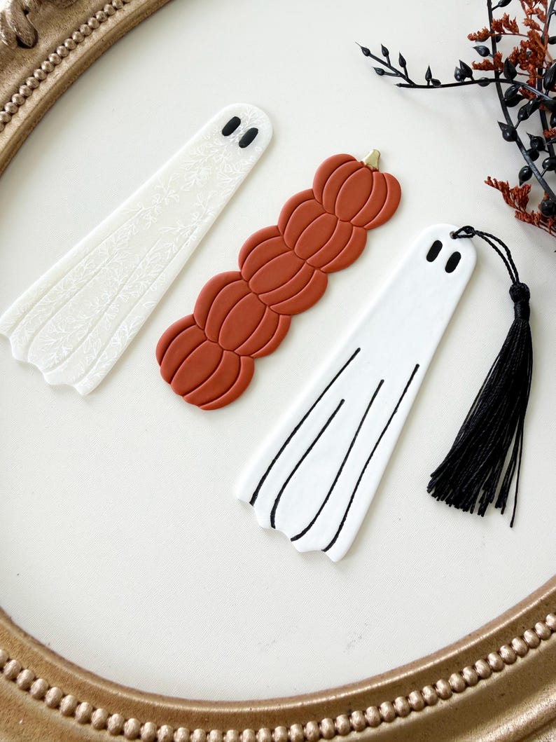 May include: Three bookmarks including a white ghost with a floral pattern, a stack of orange pumpkins, and a white ghost with black line details and a black tassel. Each bookmark features two black oval eyes at the top for a ghost-like appearance.