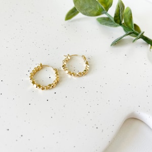 May include: A pair of gold hoop earrings with a beaded design. The earrings are on a white speckled surface.