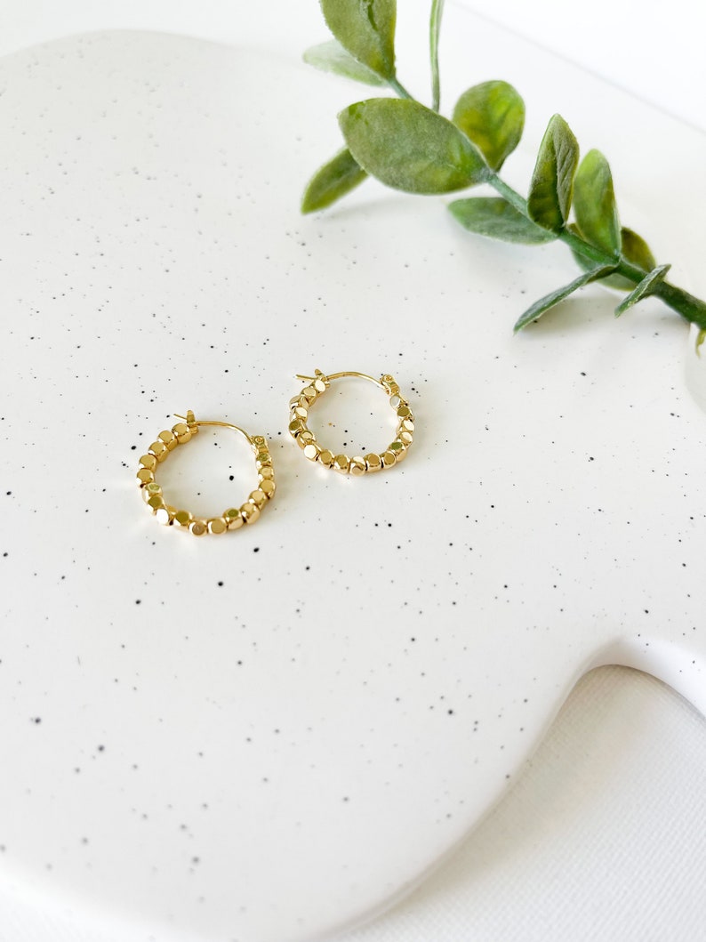 May include: A pair of gold hoop earrings with a beaded design. The earrings are on a white speckled surface.