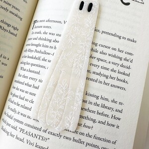 May include: A translucent, ghost-shaped bookmark featuring a delicate white botanical line art pattern and two solid black oval eyes. The bookmark is positioned vertically on an open book page with printed text, resting against the paper surface.