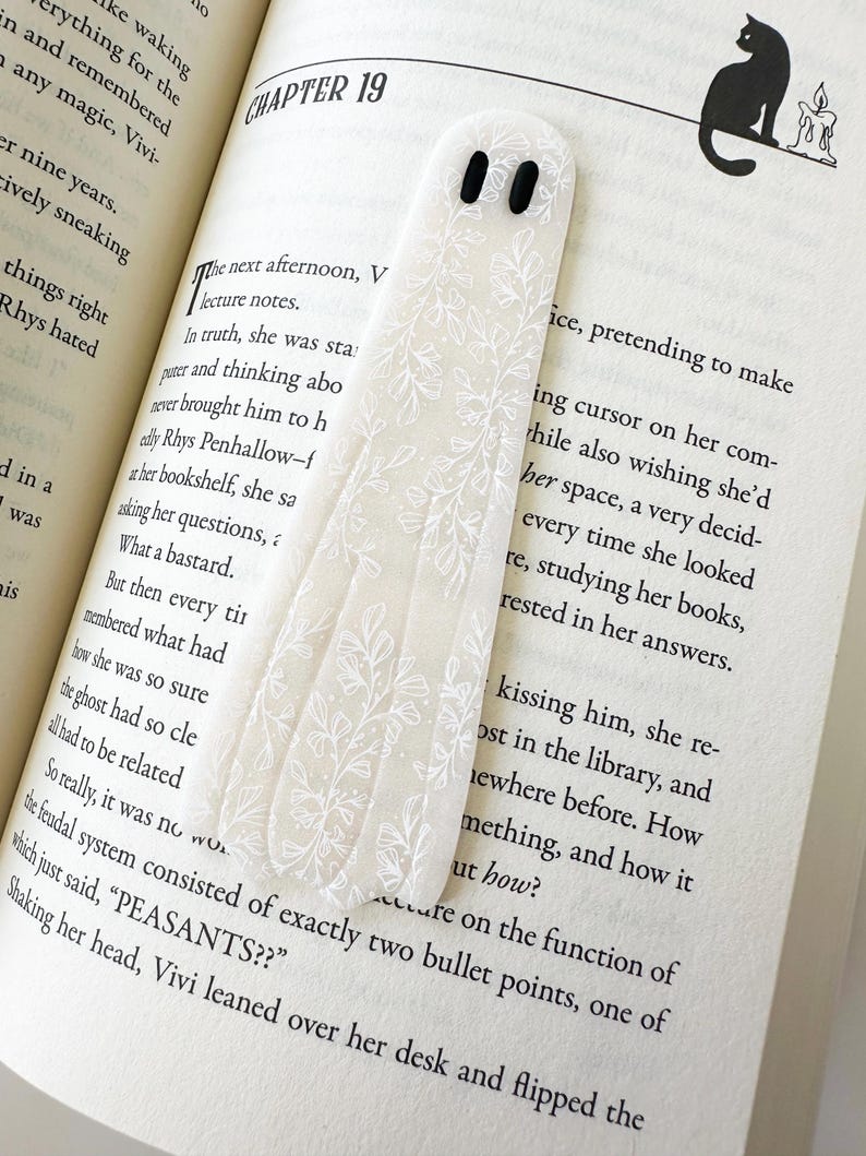 May include: A translucent, ghost-shaped bookmark featuring a delicate white botanical line art pattern and two solid black oval eyes. The bookmark is positioned vertically on an open book page with printed text, resting against the paper surface.