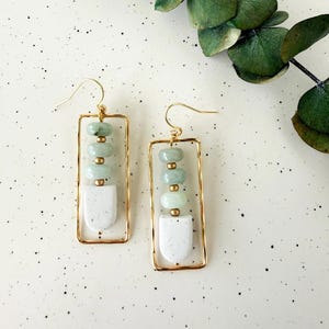May include: A pair of gold-colored rectangular earrings. Each earring features a series of light green beads, small gold-colored beads, and a white, speckled, shield-shaped element. The earrings are displayed against a speckled white background.