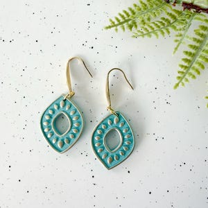 May include: A pair of turquoise teardrop earrings with a gold-colored hook. The earrings have a textured design with a central oval and surrounding petal shapes. The earrings are displayed on a white surface with a sprig of greenery.