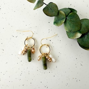 May include: Gold-tone dangle earrings with a hoop design. Each earring features a green, cylindrical bead, small gold beads, and white beads. The earrings are displayed on a speckled white surface with green foliage.
