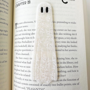 May include: A translucent, ghost-shaped bookmark featuring a white botanical line art pattern and two solid black oval eyes. The bookmark is placed vertically on an open book page that displays printed text and a small illustration of a cat.