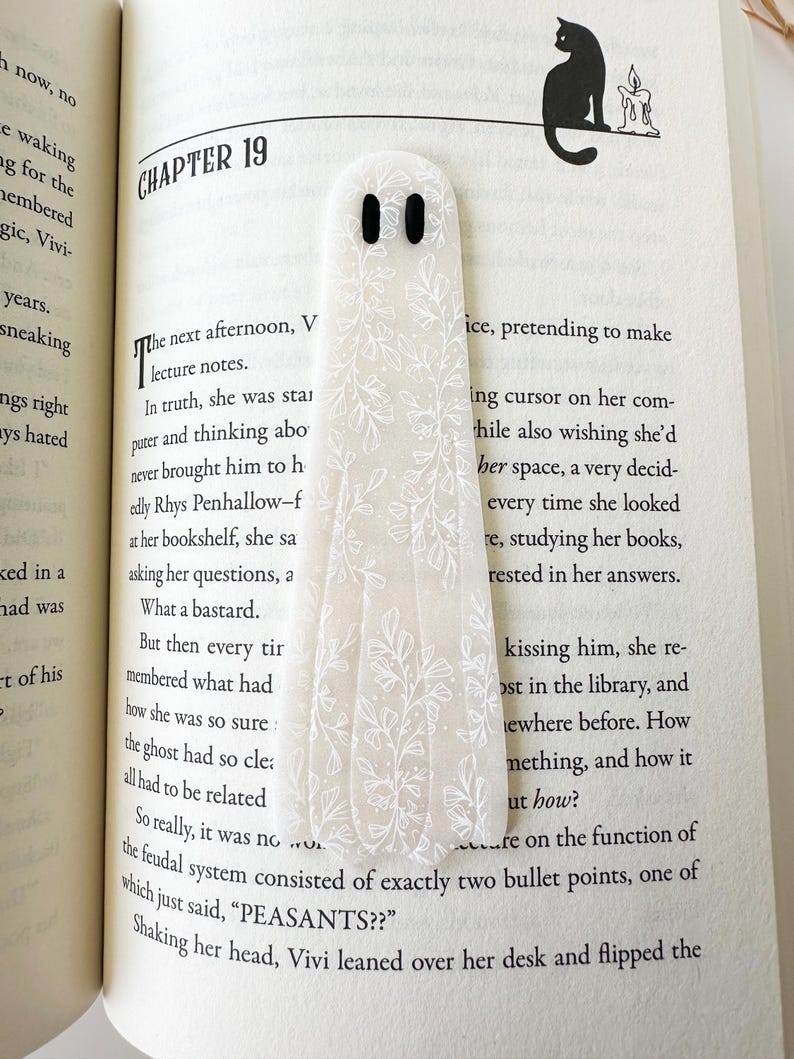 May include: A translucent, ghost-shaped bookmark featuring a white botanical line art pattern and two solid black oval eyes. The bookmark is placed vertically on an open book page that displays printed text and a small illustration of a cat.