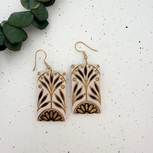 May include: A pair of rectangular earrings with a cream-colored base and gold and brown floral design. The earrings have gold-colored hooks. The design features a stylized tree and sunburst pattern.