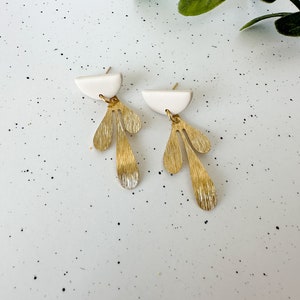 May include: A pair of gold and white earrings with a geometric design. The earrings feature a white half-circle top with a gold dangle that resembles a stylized leaf.