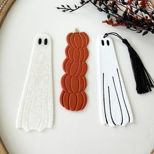 May include: Three bookmarks: one white ghost with a delicate white floral pattern, one stack of five burnt orange pumpkins with a gold stem, and one white ghost with black line details and a black tassel attached to the top.