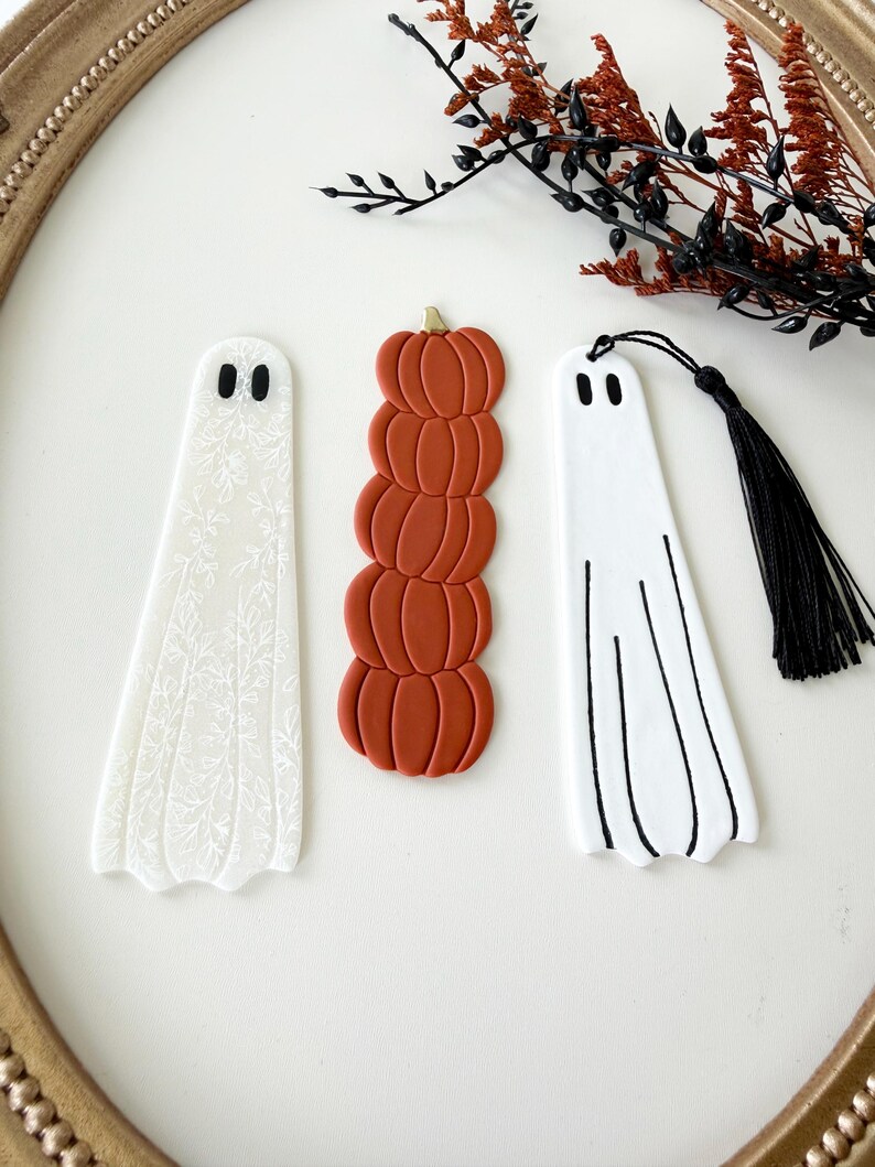 May include: Three bookmarks: one white ghost with a delicate white floral pattern, one stack of five burnt orange pumpkins with a gold stem, and one white ghost with black line details and a black tassel attached to the top.