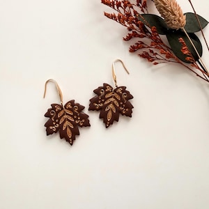 May include: A pair of dark brown maple leaf-shaped earrings featuring intricate gold-colored filigree detailing on the surface. Each earring is attached to a simple gold-toned metal hook, creating a cohesive set of jewelry with a matte finish.