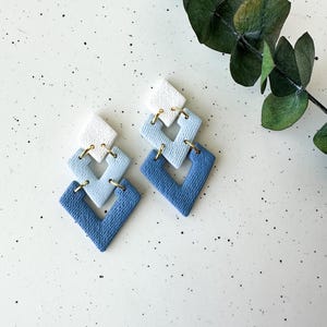 May include: A pair of blue and white geometric earrings with gold accents. The earrings are made of polymer clay and have a layered design.