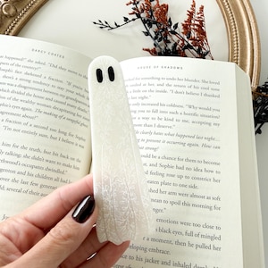 May include: A white ghost-shaped bookmark featuring a delicate, repeating white floral pattern and two solid black oval eyes. The bookmark is positioned vertically against the open pages of a book, showcasing its smooth, flat, and elongated form.