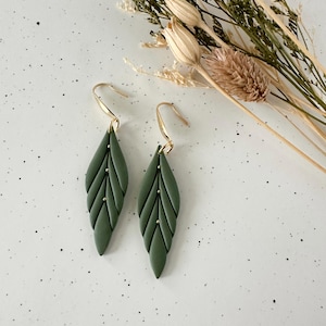May include: A pair of olive green leaf-shaped earrings with gold-colored hooks and small gold-colored accents. The earrings are displayed on a speckled white surface, with dried flowers in the background.