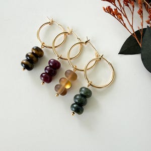 May include: Four gold hoop earrings, each featuring a trio of dangling beads. The beads include brown tiger's eye, deep burgundy, translucent amber, and dark green stone, all finished with a small gold bead at the bottom of each earring strand.
