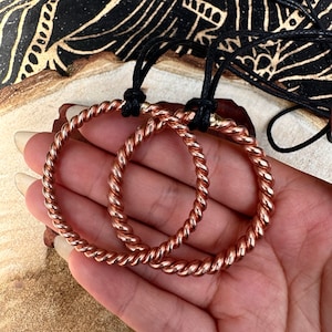 Op de afbeelding: Twee koperen draad armbanden met een gedraaid ontwerp. De armbanden zijn op een hand en zijn verbonden door een zwart koord.