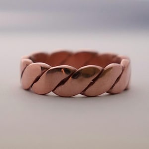 May include: A copper-colored ring with a braided design. The ring has a continuous pattern of interwoven strands, creating a textured surface. The ring is a warm, reddish-brown color.