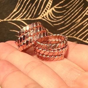 Puede incluir: Un conjunto de tres anillos de cobre con un diseño trenzado. Los anillos están apilados uno encima del otro y son de un color marrón rojizo cálido.