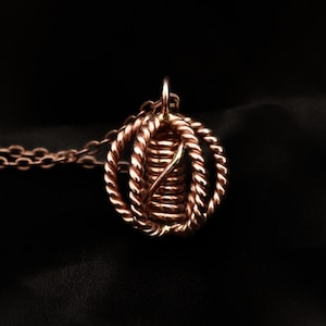 Puede incluir: Un collar con colgante de color cobre. El colgante presenta un diseño en espiral dentro de un marco de alambre retorcido. La cadena del collar también es de color cobre. El fondo es negro liso.