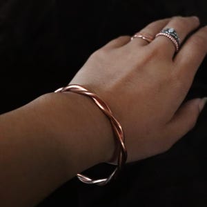 Può includere: Un braccialetto in filo di rame con un design intrecciato. Il braccialetto è indossato al polso.