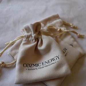 May include: Close-up of two beige drawstring pouches with the text "COZMIC ENERGY • ELEVATE YOUR FREQUENCY" printed in black. The pouches are made of a soft material and have a drawstring closure.