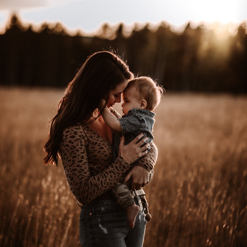 Peut inclure: Une femme portant un chemisier l&eacute;opard brun et noir tient un jeune enfant dans ses bras. L'enfant porte une chemise en jean bleu et un pantalon camouflage. Ils se tiennent dans un champ d'herbe haute.