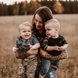 Peut inclure: Une femme portant une chemise &agrave; imprim&eacute; l&eacute;opard tient deux tout-petits dans ses bras. Le tout-petit &agrave; gauche porte une chemise en jean bleu et un pantalon camouflage. Le tout-petit &agrave; droite porte un t-shirt noir &agrave; motif camouflage et un jean bleu.