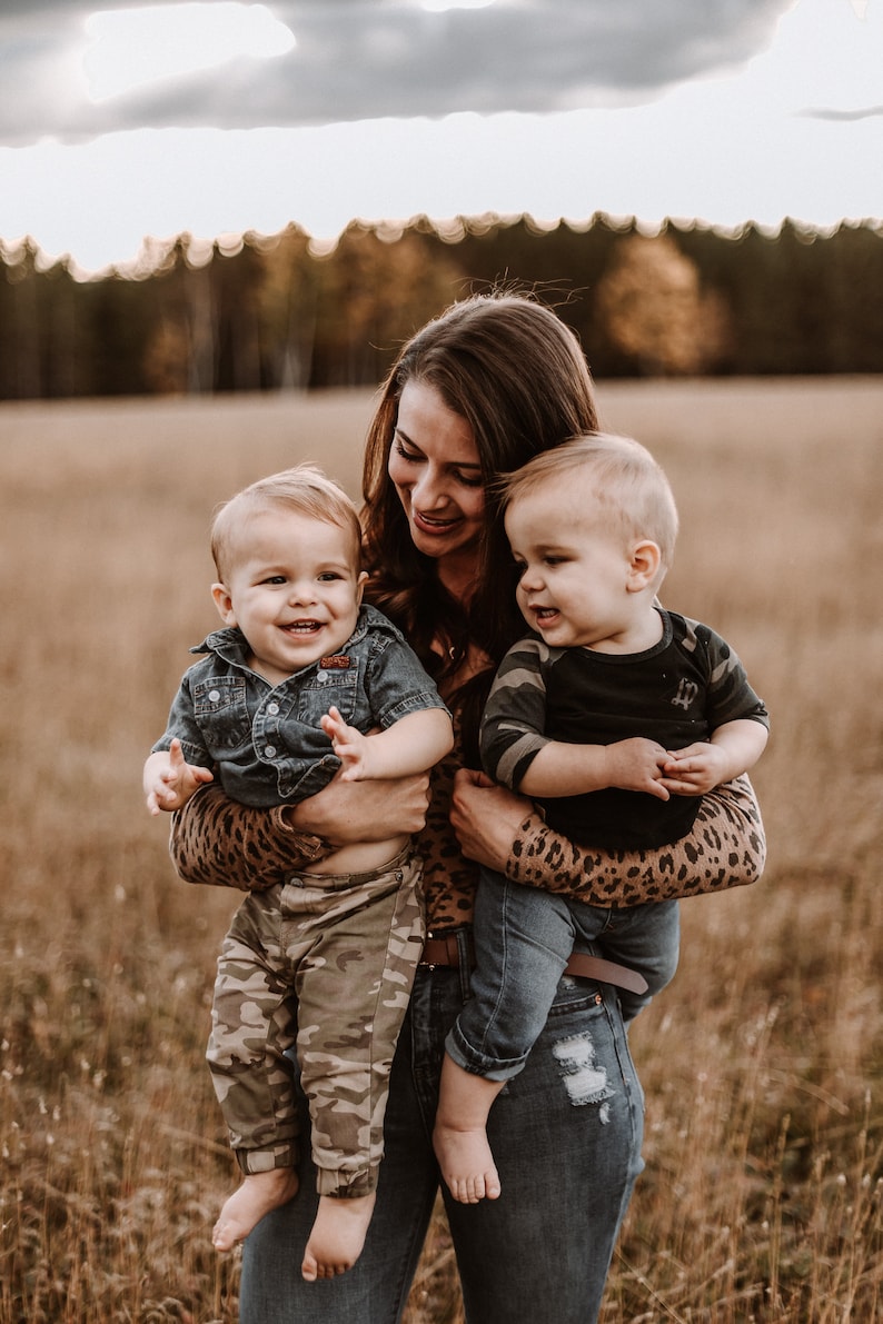 Peut inclure: Une femme portant une chemise &agrave; imprim&eacute; l&eacute;opard tient deux tout-petits dans ses bras. Le tout-petit &agrave; gauche porte une chemise en jean bleu et un pantalon camouflage. Le tout-petit &agrave; droite porte un t-shirt noir &agrave; motif camouflage et un jean bleu.