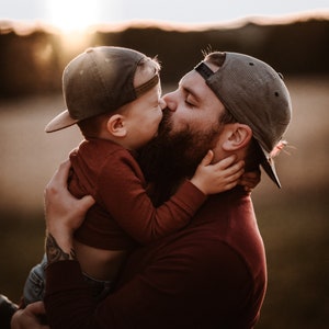 Peut inclure: Un p&egrave;re et son fils, tous deux v&ecirc;tus de chemises rouges et de casquettes de baseball, s'embrassent et se serrent dans leurs bras. Le p&egrave;re a une barbe et un tatouage sur le bras. L'arri&egrave;re-plan est un champ avec un coucher de soleil au loin.