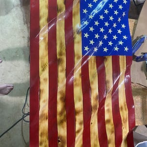 May include: Wooden American flag with a distressed finish. The flag features red and yellow stripes, a blue field with white stars, and a wavy design. The wood grain is visible, adding texture to the patriotic artwork.