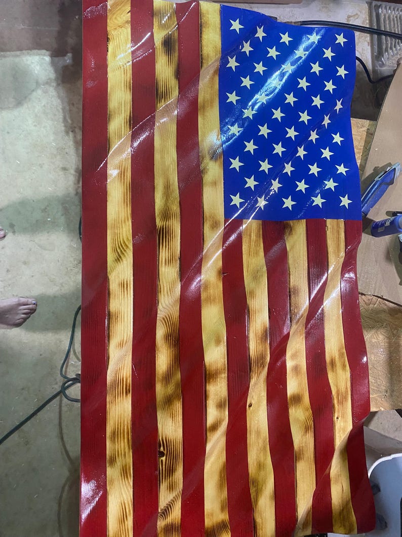 May include: Wooden American flag with a distressed finish. The flag features red and yellow stripes, a blue field with white stars, and a wavy design. The wood grain is visible, adding texture to the patriotic artwork.